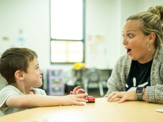 GVL A young boy and a woman sit at a table. The boy smiles and holds a toy car, while the woman looks at him with a surprised and excited expression. They appear to be interacting joyfully during ABA therapy at Project Hope Foundation.