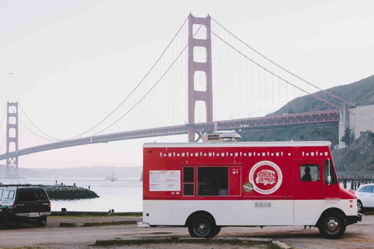 GVL A red food truck is parked near the water in San Francisco with the Golden Gate Bridge in the background on a cloudy day. The truck has a white menu board, a logo on the side, and serves as a great example for Food Truck Rentals. A black car is parked nearby.