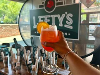GVL A person holds a bright orange cocktail garnished with an orange slice in front of a Lefty’s West End Tavern sign inside a modern bar, capturing the debut of this vibrant drink for the Weekly Dish. Tables and other drink items are visible in the background.