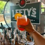 GVL A hand holds up a bright orange-red cocktail with an orange slice garnish in front of a sign that says “Lefty’s.” Several drink tap handles are visible in the background at Lefty’s West End Tavern, perfect for your next Weekly Dish debut.