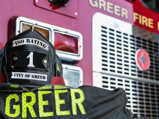 GVL A firefighter wearing gear labeled “GREER” stands before a red Greer Fire Department truck, its helmet reading “ISO Rating 1, City of Greer”—highlighting the city’s equipment purchase approved by the City Council.
