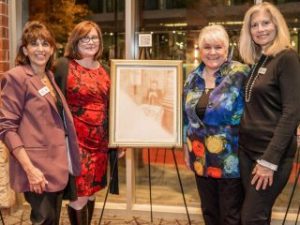 GVL Four women stand indoors, smiling beside a framed, faded portrait on an easel. Dressed in business or semi-formal attire, they gather at a Greenville Jewish Film Festival event, with large windows and nighttime city lights in the background.