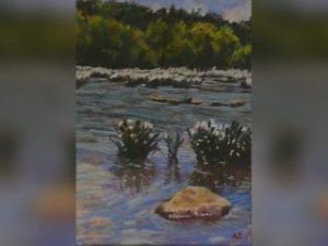GVL A painting of a calm river with rocks and small plants in the shallow water, featured in the Taking a Hike exhibition at Simpsonville Arts Center, with dense green trees along the opposite riverbank under a partly cloudy sky.