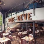 GVL A modern restaurant interior with wooden tables and chairs, large windows, and a mezzanine level. The wall features large marquee letters spelling MOSE DIVE. Perfect for hosting a charity brunch to support Texas flood victims; the space is empty and well-lit.