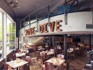 GVL A modern restaurant interior with wooden tables and chairs, large windows, and a mezzanine level. Illuminated marquee letters spelling NOSE DIVE are mounted above the main dining area—part of the Table 301 group.