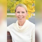 GVL A woman with blonde hair in a low bun, wearing a white cowl-neck sweater, sits on a bench outdoors amid bright yellow foliage in Greenville, smiling at the camera as if shes just finished reading a gripping novel.