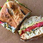 GVL A toasted tomato sandwich cut in half, filled with layers of lettuce, tomato, and possibly cheese or avocado, displayed on brown parchment paper.