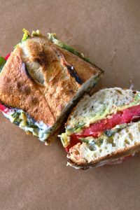 GVL A crusty Tomato Sandwich cut in half, filled with layers of tomato, avocado, leafy greens, and a creamy spread, placed on brown parchment paper.