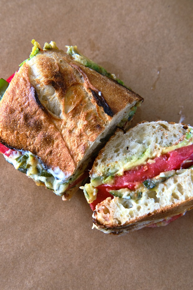 GVL A crusty Tomato Sandwich cut in half, filled with layers of tomato, avocado, leafy greens, and a creamy spread, placed on brown parchment paper.