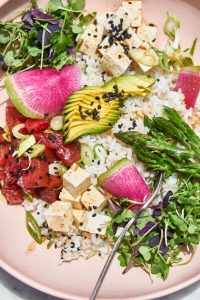 GVL A vibrant vegetarian poke bowl with white rice, diced tofu, avocado, seared tuna, watermelon radish, asparagus, microgreens, black sesame seeds, and scallions on a pink plate, with a fork resting inside.