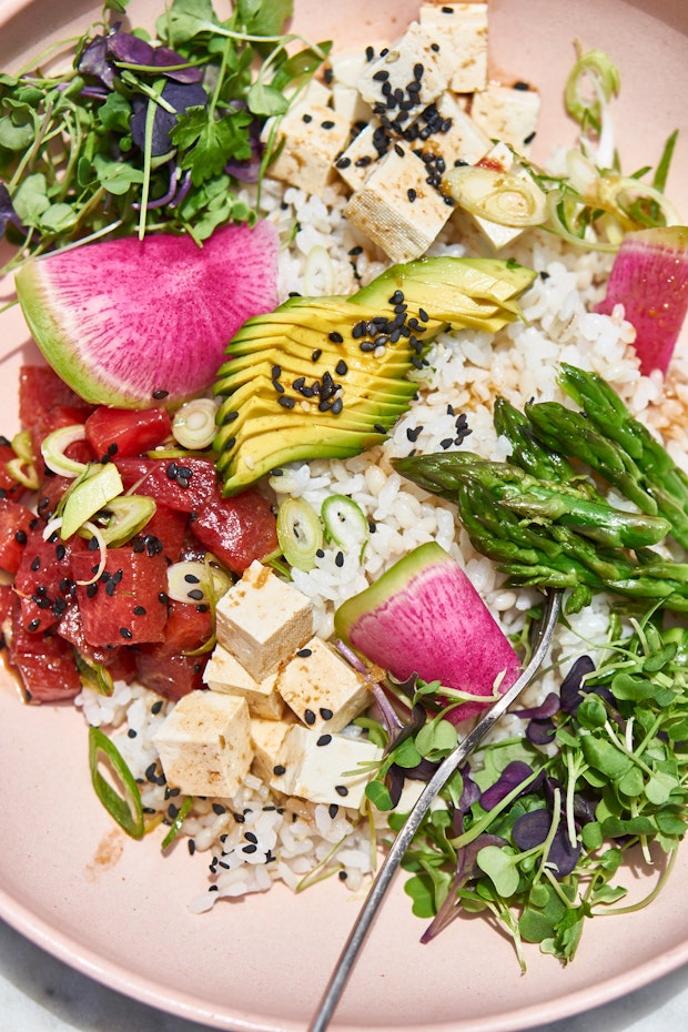 GVL A vibrant vegetarian poke bowl with white rice, diced tofu, avocado, seared tuna, watermelon radish, asparagus, microgreens, black sesame seeds, and scallions on a pink plate, with a fork resting inside.
