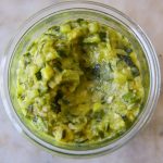 GVL A glass jar filled with a chunky green and yellow mixture, likely a freshly made Zucchini Butter or savory condiment with herbs and vegetables, shown from above on a light surface.