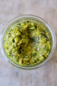 GVL A glass jar filled with chunky, green Zucchini Butter sits on a light, marbled surface. The spread appears to have pieces of avocado and green herbs mixed in.